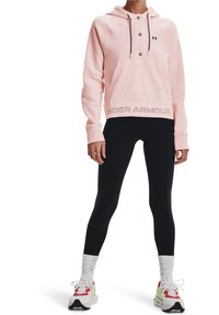 Sudadera con capucha de color rosa claro y diseño con botones, bolsillo frontal y "UNDER ARMOUR" impreso en gris en el dobladillo. Combinada con leggings negros y zapatillas deportivas.