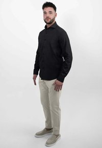 Zwart overhemd met een slim fit, gecombineerd met lichtbeige broeken en crèmekleurige instappers, staand tegen een effen achtergrond.
