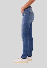 Schmale blaue Jeans in mittlerem Waschton mit fünf Taschen. Mit gelben Nähten an der Gesäßtasche. Getragen mit weißen Turnschuhen.