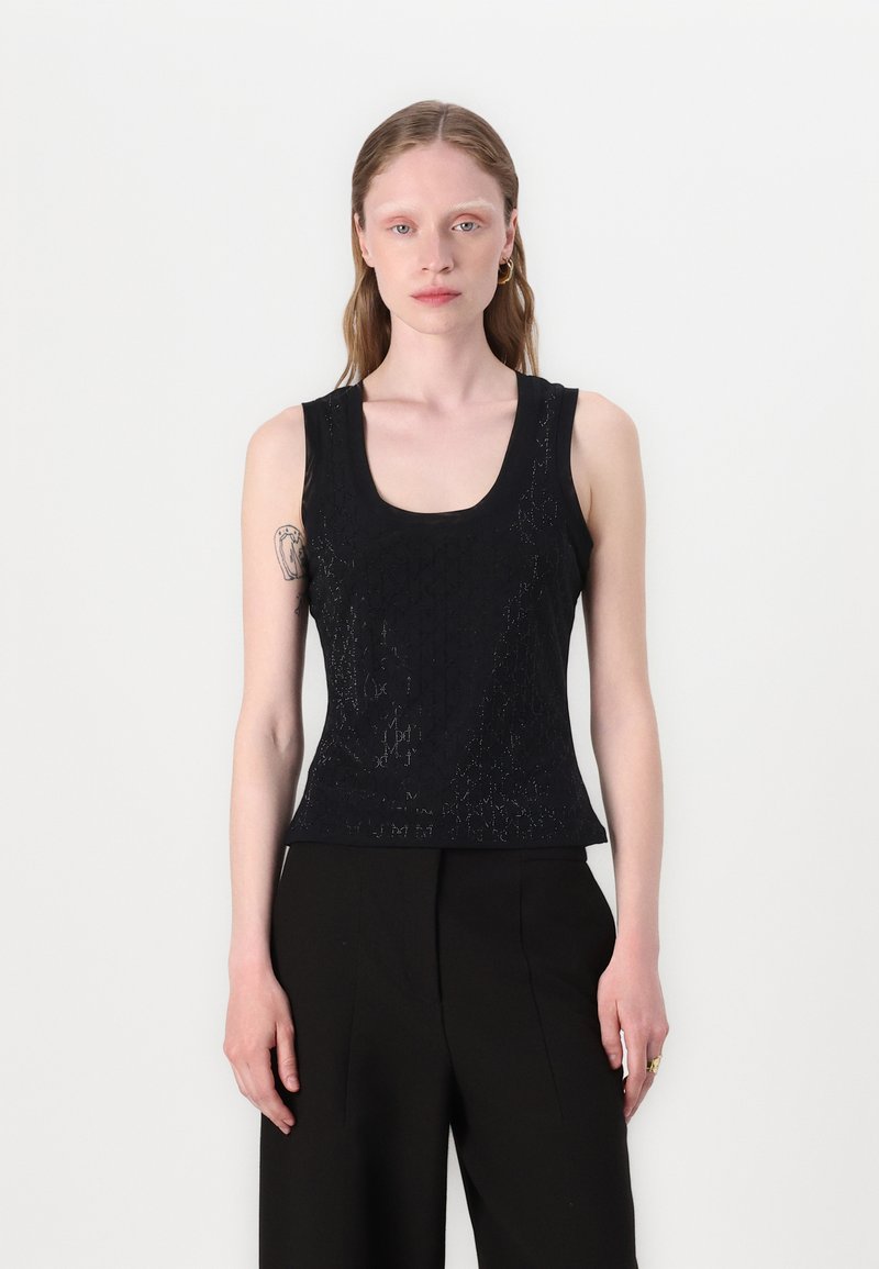 Femme à la peau claire et aux longs cheveux, portant un haut noir sans manches texturé et un pantalon noir, debout devant un fond clair uni.