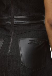 Μαύρο denim σύνολο με γυαλιστερό μαύρο δερμάτινο περίγραμμα και τσέπη με κεντημένο "Karl" κοντά στο ορατό χέρι και στο κεντρικό φερμουάρ.