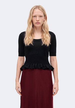Femme blonde aux cheveux ondulés porte un haut noir à manches courtes avec un ourlet à volants et une jupe rouge foncé, debout sur un fond clair.