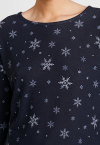 Haut à manches longues bleu marine avec un motif de flocons de neige en blanc et bleu clair, confectionné dans un tissu doux et texturé avec un col arrondi.