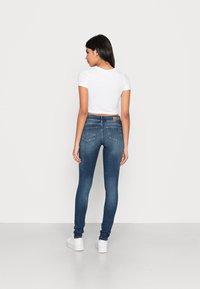 Jeans skinny em denim azul escuro, com uma cintura de altura média, dois bolsos traseiros e um rótulo de couro castanho na cintura. Acompanha ténis brancos.