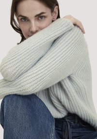 Frau mit hellblauem Strickpullover und blauen Jeans, die teilweise ihr Gesicht mit ihrem Arm bedeckt und in die Kamera schaut.