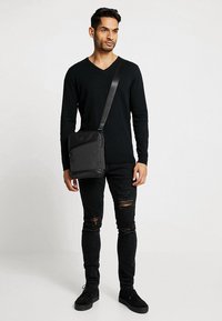 Borsa a tracolla nera con tessuto testurizzato, tasca con zip diagonale e tracolla regolabile. Il modello indossa un dolcevita nero e jeans strappati.