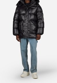 Muž v černé lesklé péřovce s kapucí, světle modrých džínách a bílých teniskách, stojící na jednobarevném bílém pozadí.