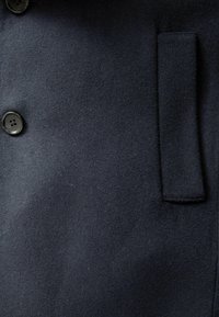Tejido de lana azul marino con una textura suave, que cuenta con dos grandes botones negros y un bolsillo lateral. El diseño es simple y funcional.