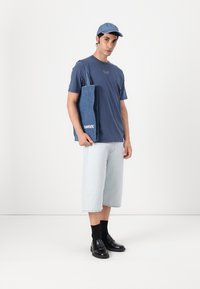 Model in a blue T-shirt, light blue denim shorts, black loafers, and a denim tote bag with "SAMSOE" logo, all against a neutral background.