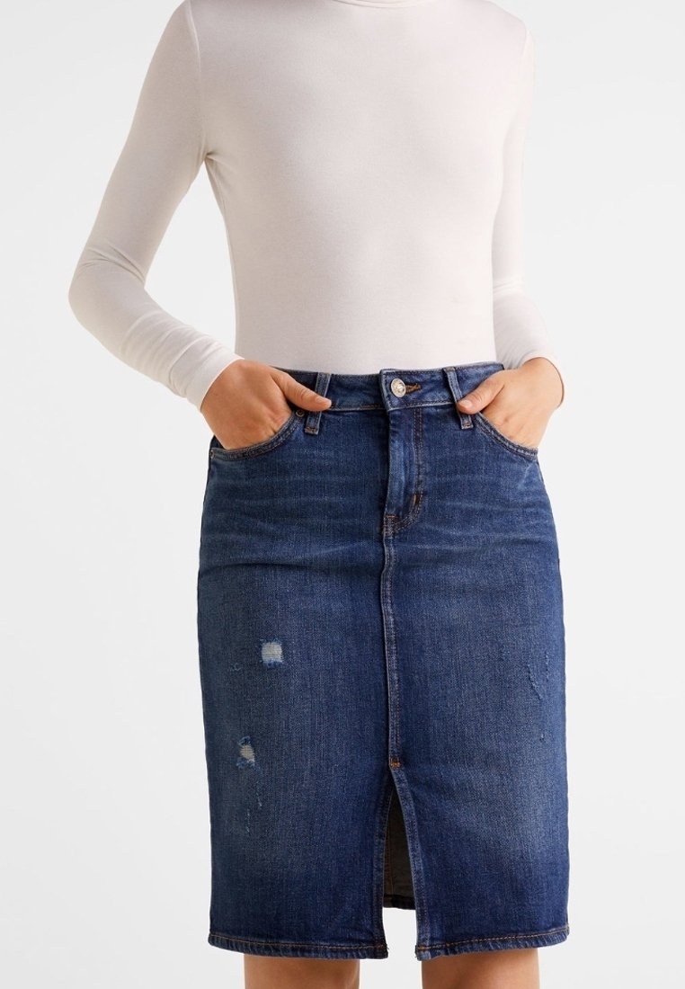 Personne portant un haut blanc ajusté à manches longues rentré dans une jupe en denim bleu foncé taille haute, longueur au genou, légèrement usée avec une fente à l'avant.
