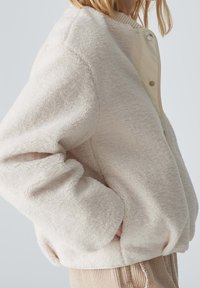 Femme portant une veste en polaire beige texturée avec des poches, associée à un pantalon en velours côtelé marron clair sur un fond uni.