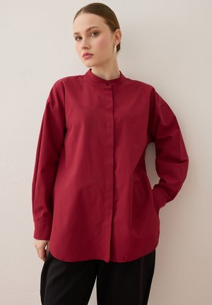 Kvinde med lys hud og tilbagebundet hår iført en langærmet rød skjorte med knapper og sorte bukser, stående mod en ensartet lys væg.