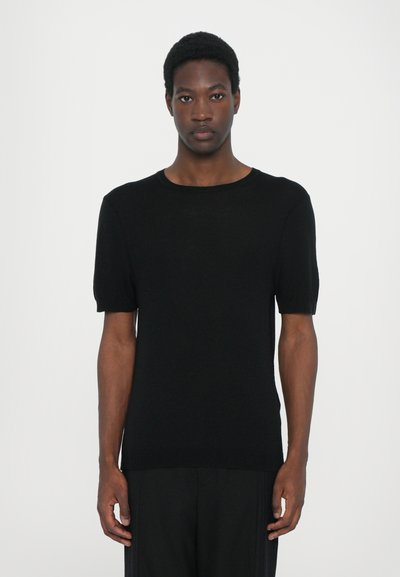 Haut tricot noir à manches courtes avec un col rond, présentant une texture lisse et une coupe ajustée. Le mannequin se tient devant un fond blanc.