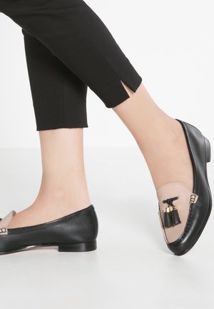 Füße in schwarzen und beigen Leder-Loafers mit Quastenakzenten, kombiniert mit schwarzen, gekürzten Hosen mit Knöchelschlitzen, stehend auf einem weißen Boden.