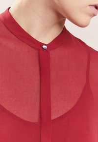 Rød gennemsigtig bluse med en rund krave, der har en central knap og præcise syninger langs knapstolpen. Glat tekstur og letvægtsstof.