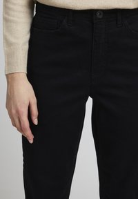 Schwarze Jeans aus Denim mit einer taillierten Passform, ausgestattet mit Fronttaschen, einem Knopfverschluss und einer glatten Textur. Sichtbare Nähte und Fugen.