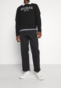 Sudadera negra con el texto blanco "GUESS EST. 1981", superpuesta sobre una camisa a rayas blanca y negra. Combinada con pantalones negros ajustados y zapatillas blancas.