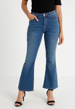 Femme portant un jean bleu taille haute évasé, des talons ouverts noirs et un haut noir, debout, une main dans la poche, sur fond blanc.