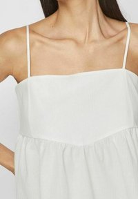 Vit tanktopp med tunna band, med en texturerad tyg, rak halsringning och en fladdrande, rynkad nederkant. Enkel, minimalistisk design.