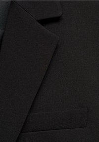 Gros plan d'une veste de costume noire montrant le revers et une poche passepoilée simple sur un tissu texturé.