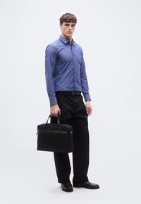 Άνδρας με μπλε πουκάμισο με κουμπιά και μαύρο παντελόνι κρατώντας μια μαύρη τσάντα, στέκεται μπροστά σε ένα απλό λευκό φόντο.
