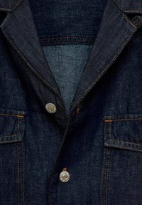 Zbliżenie na granatową kurtkę dżinsową, z kołnierzem, srebrnymi guzikami i pomarańczowym przeszyciem. Widoczne są dwie przednie kieszenie z klapkami.