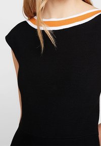 Žena v bezramínkovém černém topu s bílým a oranžovým lemem kolem výstřihu, který odhaluje krk a horní část hrudníku.