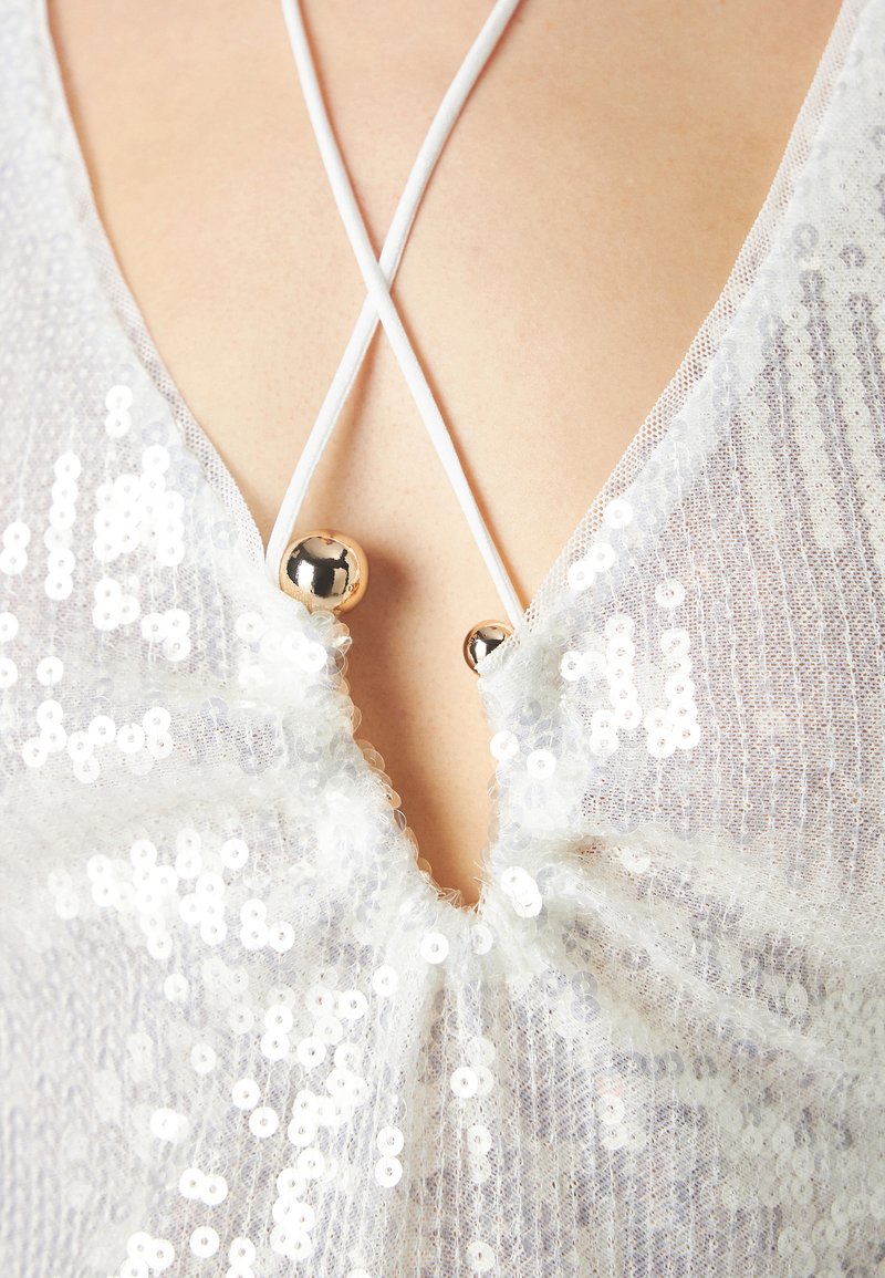 Biała przezroczysta bluzka z cekinami, głębokim dekoltem w serek i regulowanymi białymi ramiączkami, z dwoma metalowymi akcentami przy dekolcie.