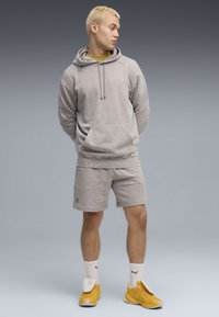 Grå bomulds-hættetrøje med en frontlomme, parret med matchende shorts. Modellen har gule sneakers på og hvide sokker, med en tekstureret finish.