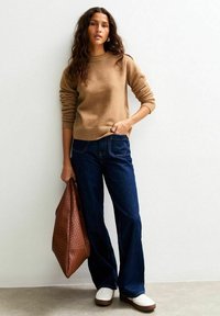 Woman with long curly hair wearing a brown sweater, dark blue jeans, white shoes, holding a large brown textured bag standing against a white wall.