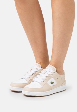 Voeten met beige en witte Lacoste-sneakers met witte veters, staand tegen een eenvoudige lichte achtergrond.