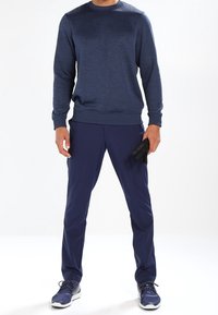 Maglione blu navy con finitura testurizzata, abbinato a pantaloni coordinati. Le sneaker presentano una suola a contrasto; guanti tenuti in una mano.