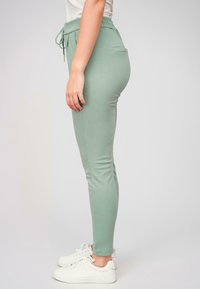 Jogging taper léger en vert, fabriqué en tissu doux, avec une taille élastique, un cordon de serrage et une poche latérale. Associé à des baskets blanches.