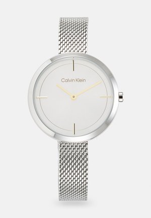 Reloj con correa de malla plateada y esfera redonda minimalista, dial gris claro, marcadores de horas dorados y el nombre de la marca "Calvin Klein" en el centro.