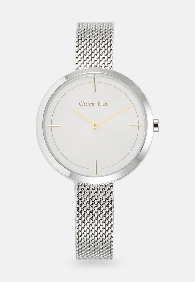 Reloj con correa de malla plateada y esfera redonda minimalista, dial gris claro, marcadores de horas dorados y el nombre de la marca "Calvin Klein" en el centro.