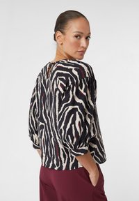 Schwarz-beige gestreiftes Zebra-Muster-Oberteil mit Puffärmeln und einem Schlüssellochdetail hinten, kombiniert mit burgunderfarbenen Hosen. Weicher, strukturierter Stoff.