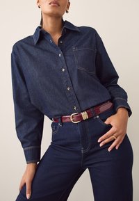 Denim-Hemd zum Zumknöpfen mit Tasche, dunkelblau und kontrastierenden Nähten, kombiniert mit hoch taillierter dunkler Jeans und einem burgunderroten Gürtel.