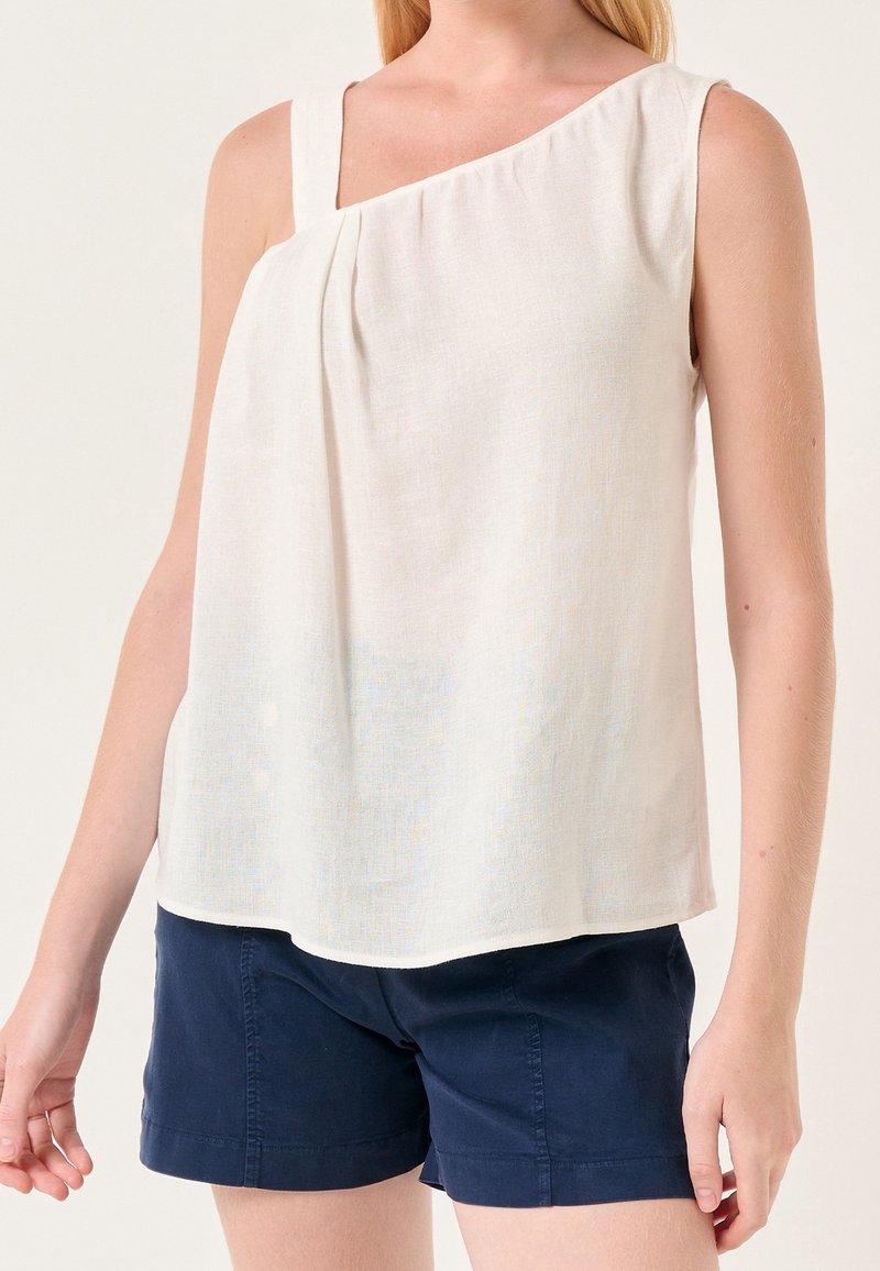Femme portant un top blanc asymétrique sans manches et un short bleu marine sur un fond clair uni.