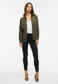 Camicia verde oliva con bottoni e due tasche sul petto, indossata sopra una canotta bianca, abbinata a jeans skinny neri e sandali neri con tacco.