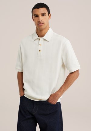 Mann mit kurzen Haaren, der ein weißes kurzärmeliges Strickpolo und dunkle Jeans trägt, mit den Händen in den Taschen vor einem einfarbigen Hintergrund.
