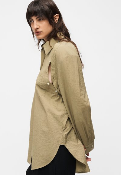 Γυναίκα με μακριά σκοτεινά μαλλιά που φοράει μια πολύ φαρδιά, μπεζ πουκαμίσα με ανοιχτές πλευρές, στέκεται μπροστά σε ένα απλό φωτεινό φόντο.