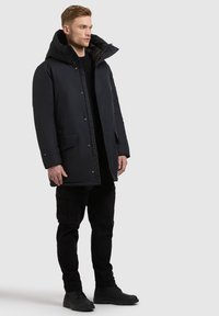 Czarna ocieplana parka z dużym kapturem, zapinana na zatrzaski, z przednimi kieszeniami, noszona na czarnym swetrze i spodniach cargo, zestawiona z czarnymi butami.