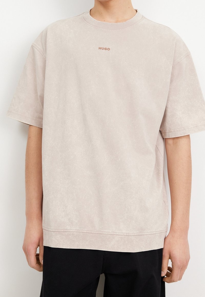 Personne portant un T-shirt beige clair à manches courtes avec le logo "HUGO" sur la poitrine et un pantalon noir sur fond blanc uni.