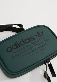 Tmavě zelený zipový pouzdro vyrobené z měkkého materiálu, s černým brandingovým textem a černým popruhem. Hladký povrch s zaoblenými okraji.