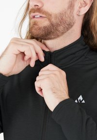 Sort zip-jakke med høj krave, fin tekstur og et hvidt logo på brystet. Hænderne justerer lynlåsen.