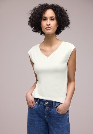 Frau mit lockigem Haar, die ein ärmelloses weißes Top und blaue Jeans trägt, mit den Händen in den Taschen vor einem einfarbigen Hintergrund stehend.