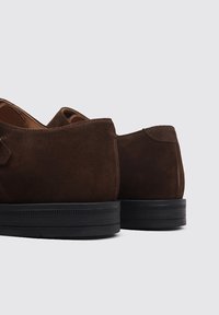Zapatos de ante marrón oscuro con una textura suave y un diseño redondeado, que cuentan con una suela de goma negra y un detalle de costura mínima.
