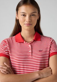 Chemise polo à rayures rouges et blanches avec un col rouge uni et des manches courtes. Présente un petit logo Levi's sur la poitrine.