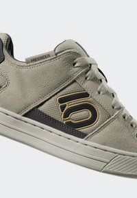 Grå mockaskor med svarta och gula accenter, platt design, snörningsstängning, texturerade sidopaneler och gummisula. "Freerider"-logotyp synlig.