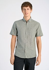 Koortriem, knoopshirt van lichtgroene textuurstof, met een borstzak en beige knopen. Gekleed met donkere broek.